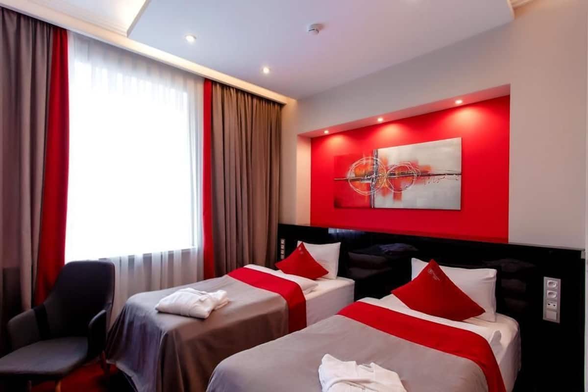 A hotel room with two beds, red and white bedding, a modern abstract painting, and a red accent wall.