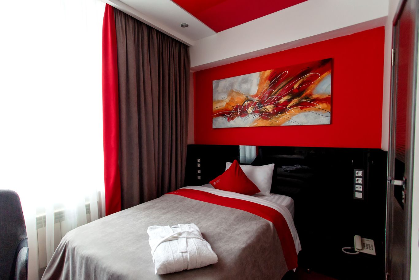 Une chambre d’hôtel avec un lit, une literie rouge et blanche, un grand tableau abstrait et un rideau rouge.