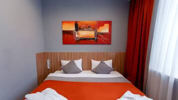 Habitación de hotel con cabecera de madera, dos almohadas, colcha naranja, cortina roja y un cuadro en la pared.
