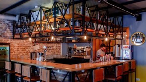Um bar com parede de tijolos, balcão de madeira e banquetas. Há uma pessoa trabalhando atrás do balcão.