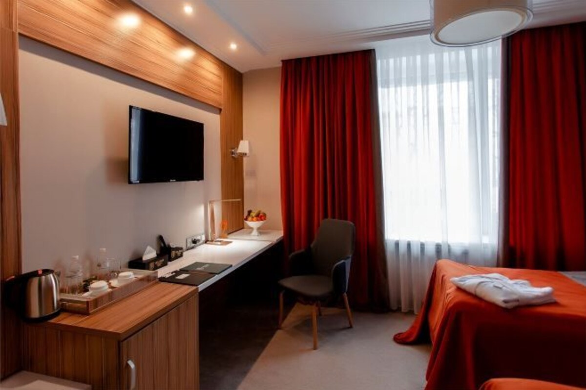 A hotel room with a wooden desk, a flat-screen TV, a bed with red curtains, and a window with sheer white curtains.