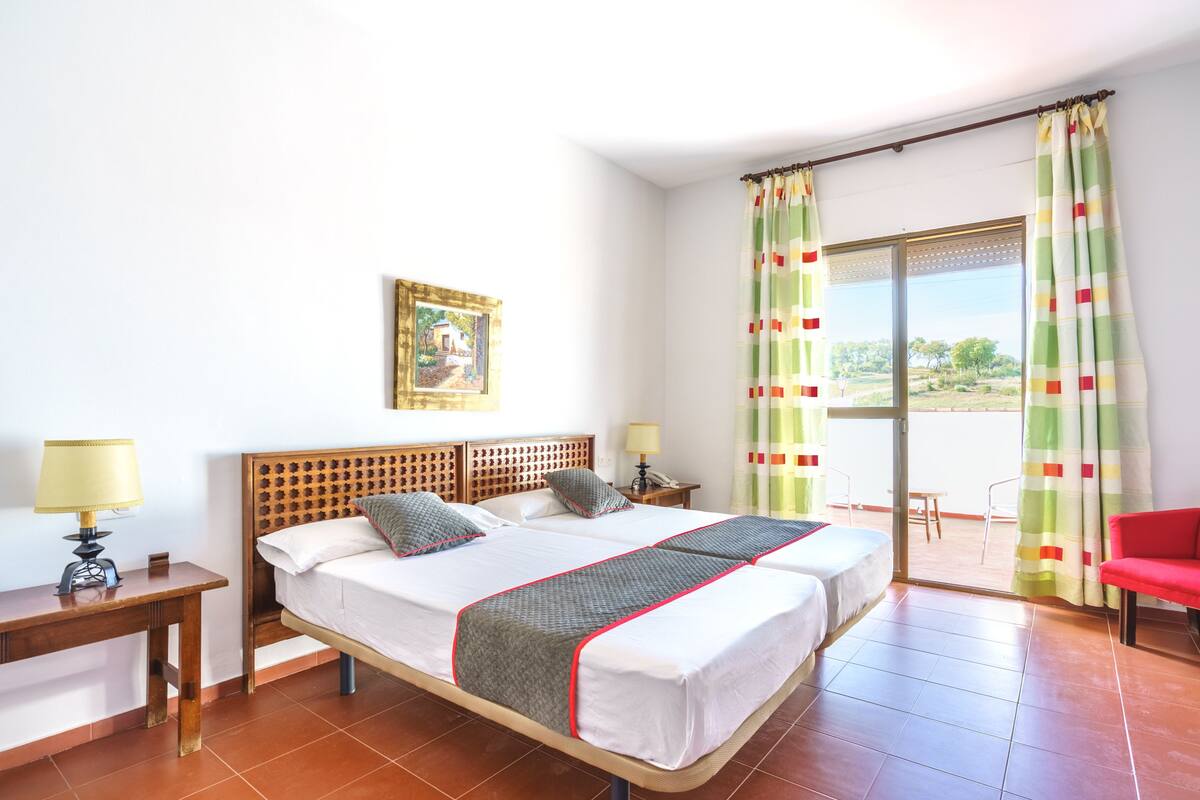 A double bed with a wooden headboard, two bedside tables with lamps, a red chair, and a view through a glass door to an outdoor area.