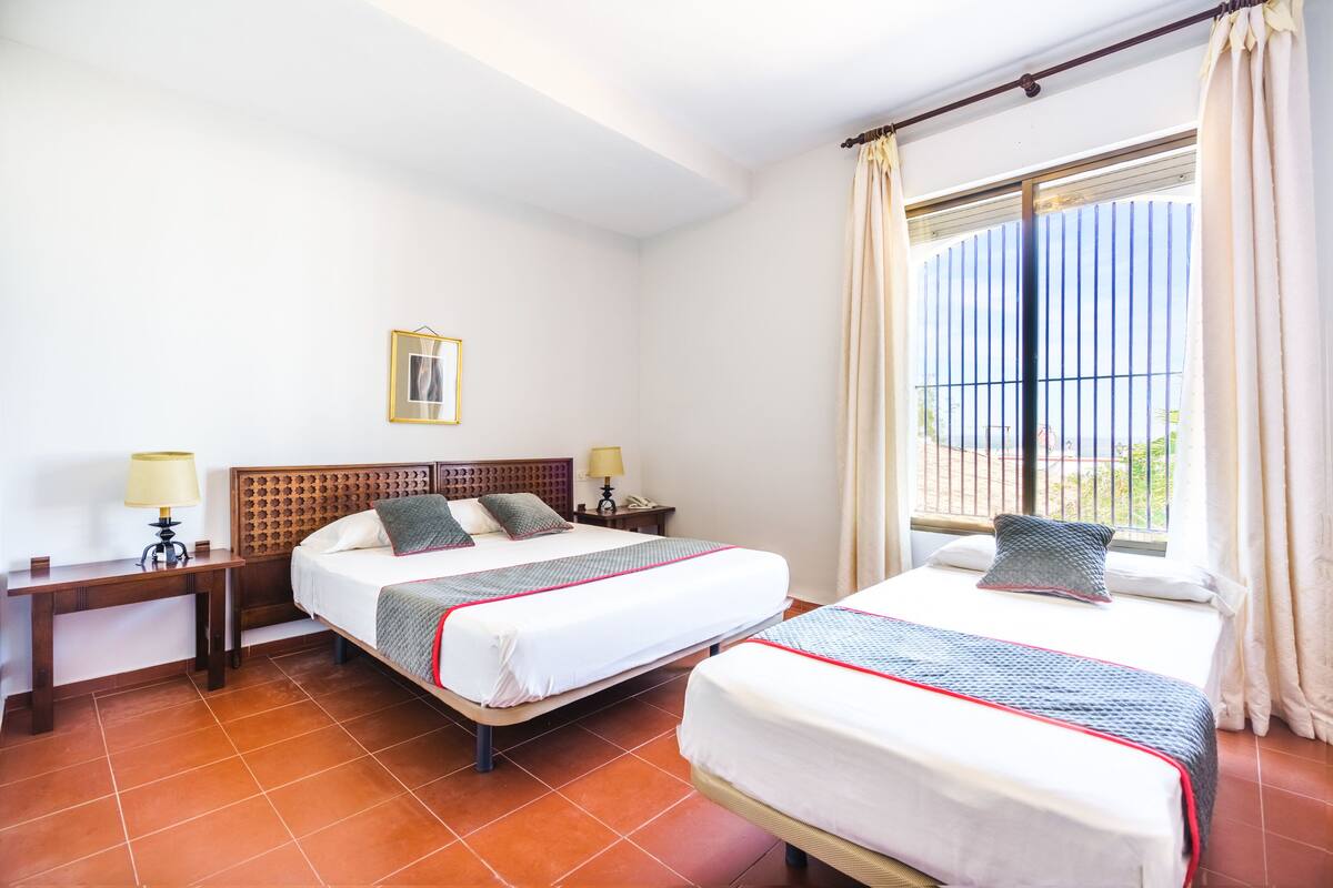 A hotel room with two beds, a wooden headboard, a nightstand with a lamp, and a window with curtains.