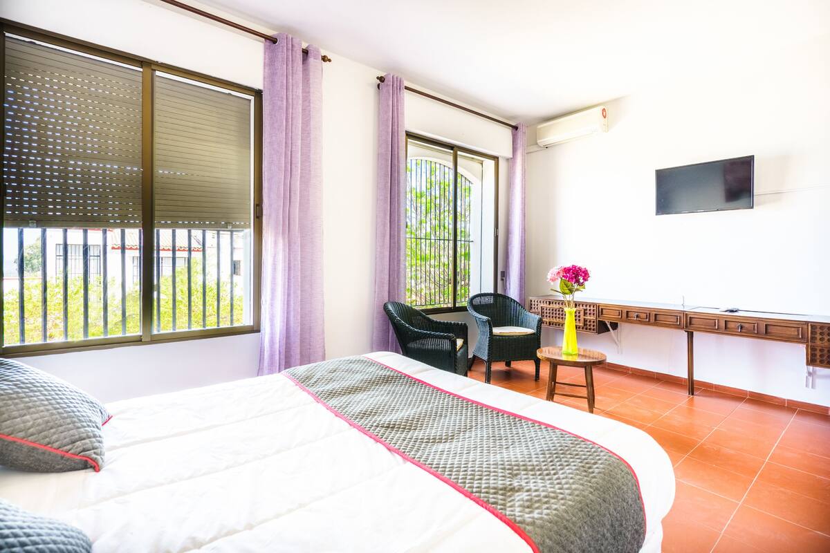 A hotel room with a bed, a chair, a small table with a vase, and a TV on the wall.
