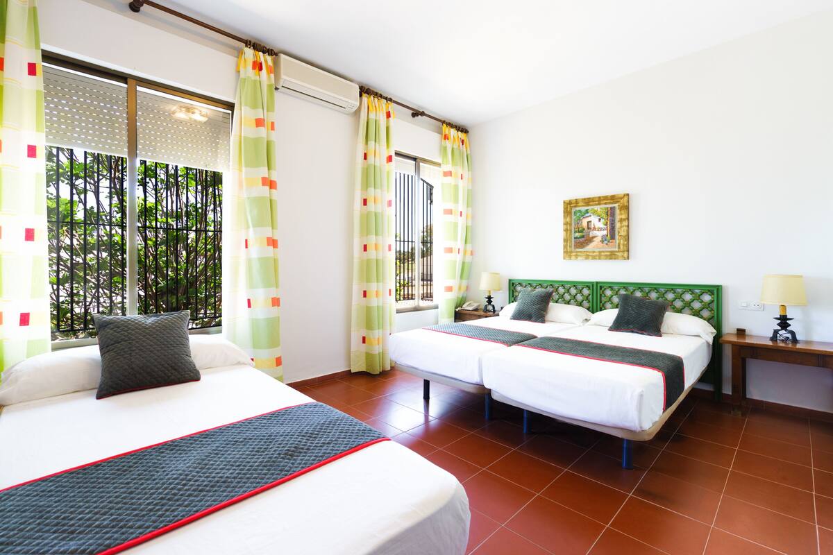 A hotel room with two beds, a green headboard, a window with curtains, a bedside table with a lamp, and a framed picture on the wall.