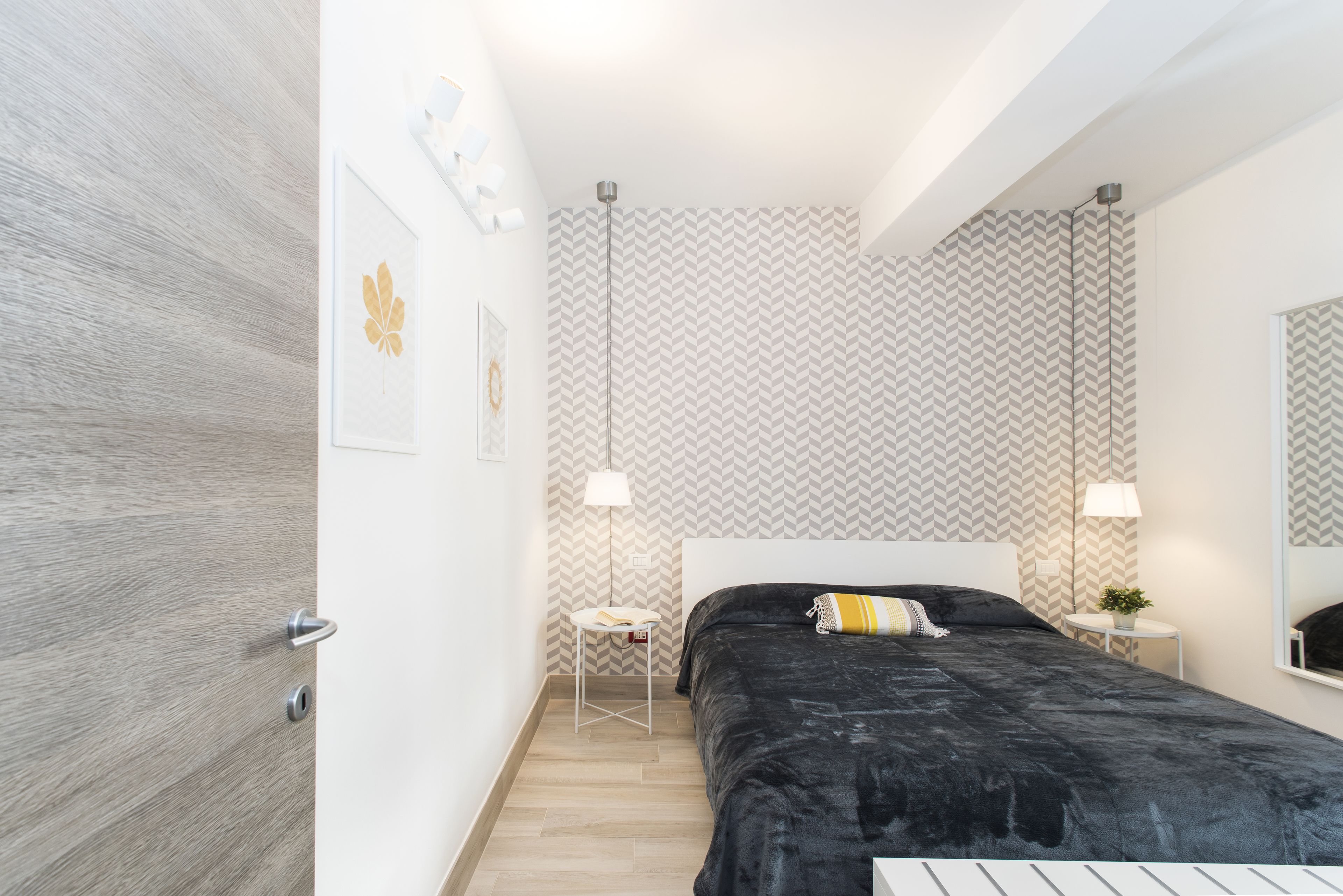 A modern bedroom with a black bed, a white bedside table, a small white stool, a wall-mounted lamp, and a mirror.