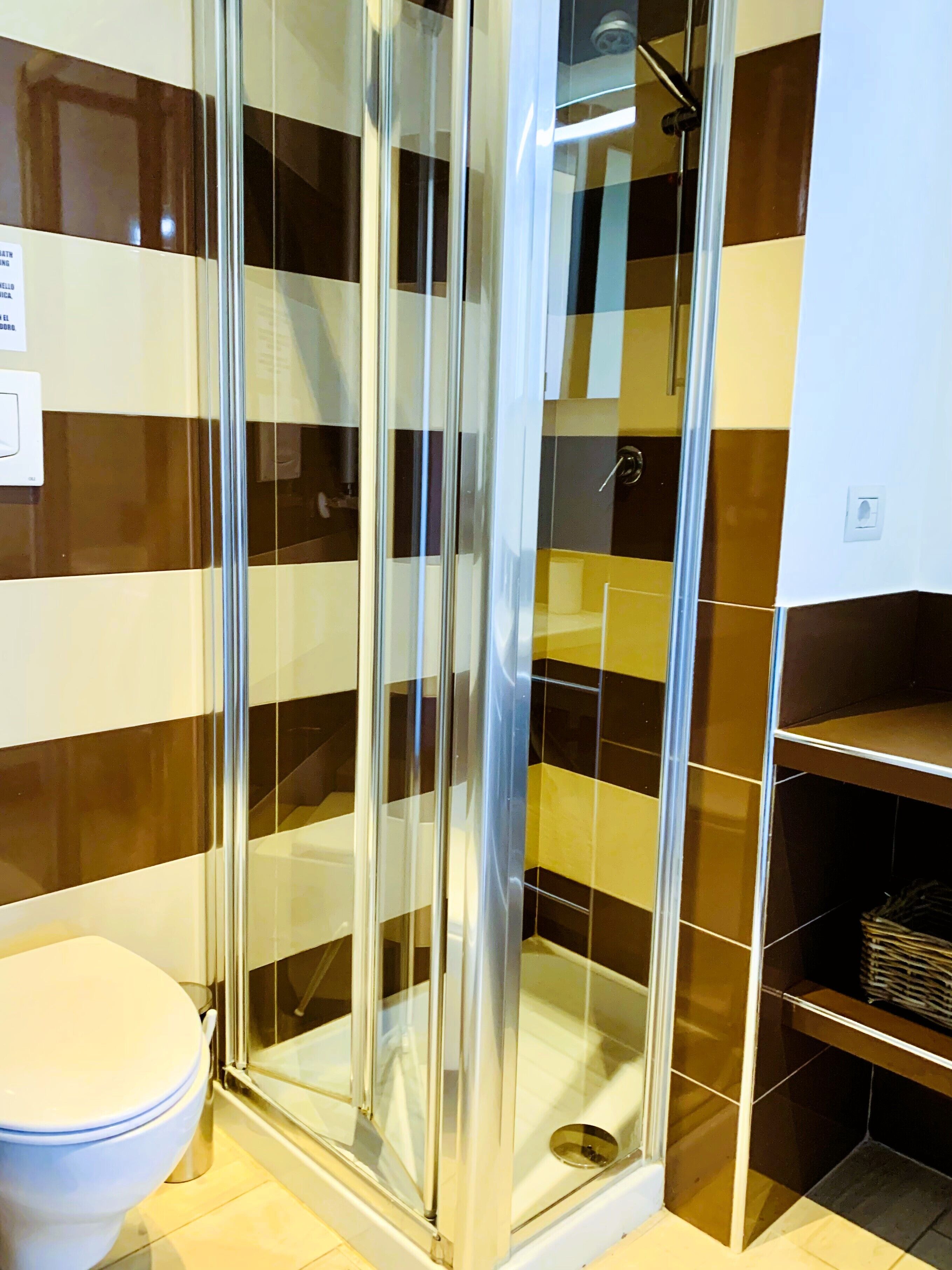 Ein Badezimmer mit einer gläsernen Duschkabine, einer weißen Toilette und einer gefliesten Wand mit braunen und beigen Mustern.