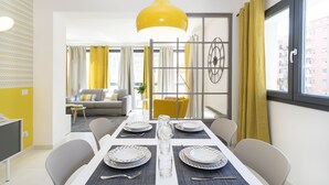 Un comedor moderno con una mesa blanca para cuatro, una lámpara colgante amarilla y vistas de la ciudad a través de grandes ventanales.