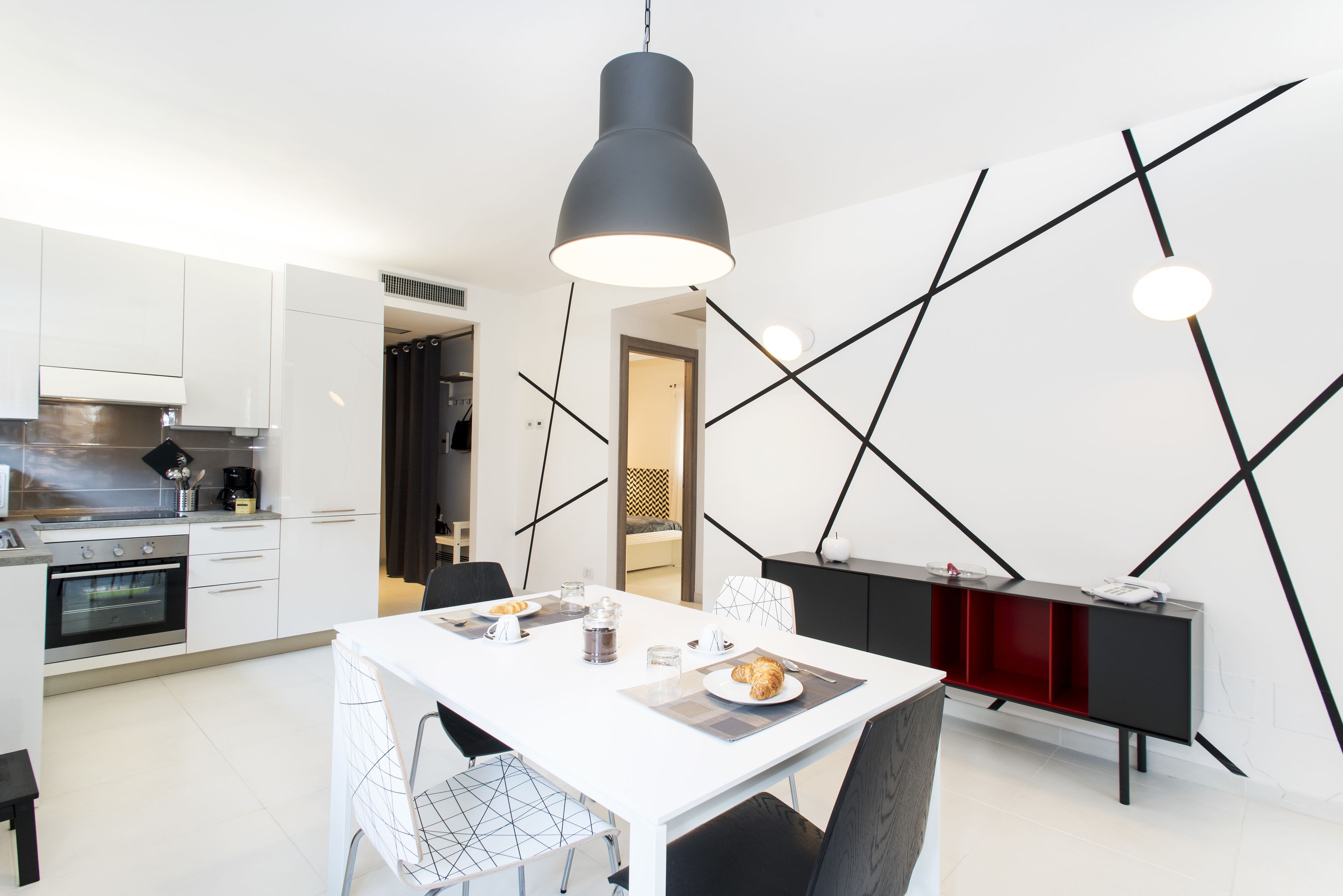 Una cucina moderna con mobili bianchi, una lampada a sospensione nera e un tavolo da pranzo apparecchiato con piatti e bicchieri.