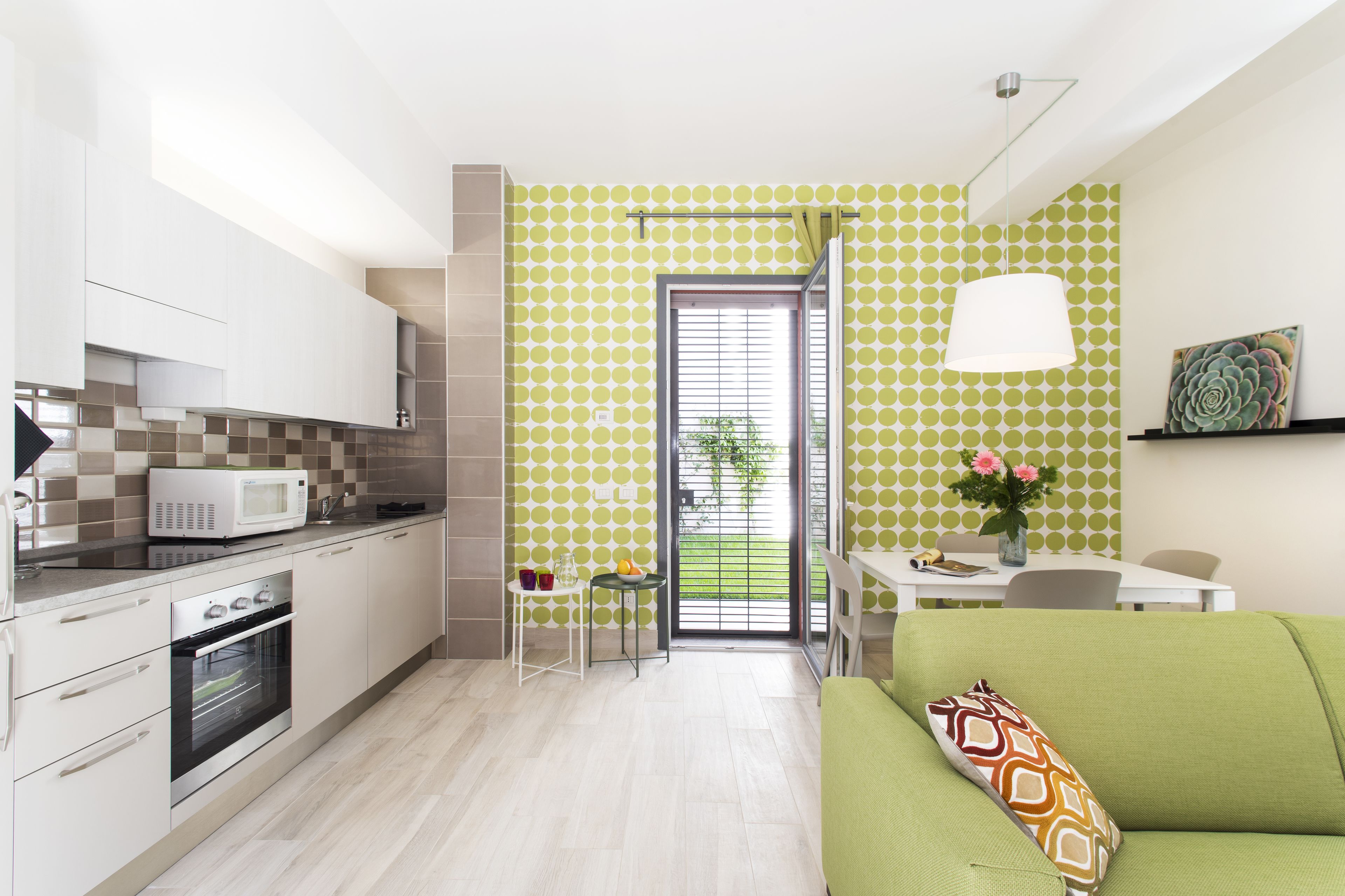 Cocina moderna con microondas, horno y armarios blancos. Hay una zona de comedor con mesa y sillas, y un sofá verde con un cojín estampado.