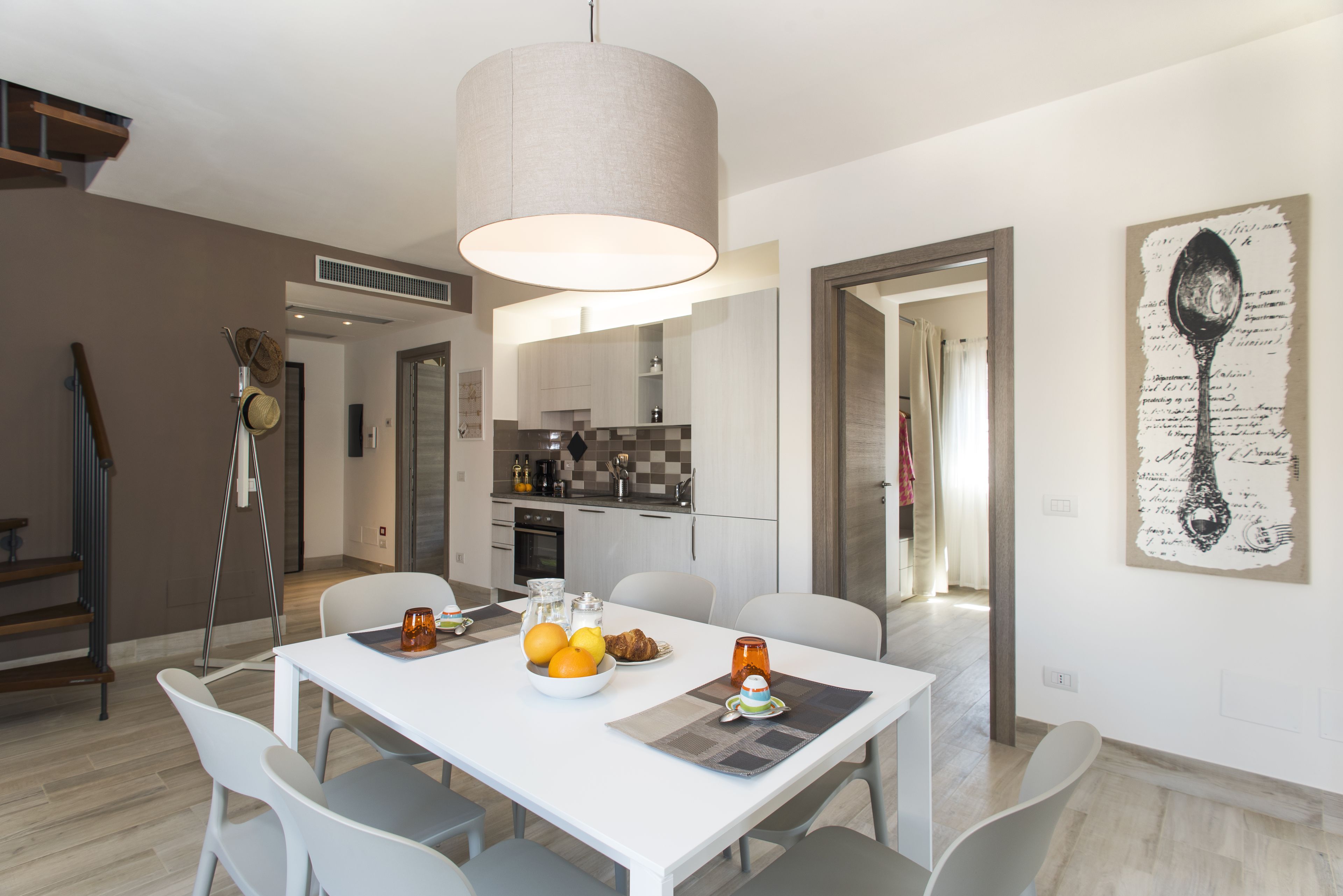 Un espace repas moderne avec une table blanche, des chaises grises et un grand luminaire suspendu. On aperçoit une cuisine en arrière-plan, avec des meubles blancs et un pare-feu à carreaux.