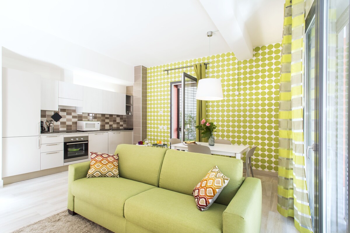 A modern living room with a green sofa, a kitchen with white cabinets, and a patterned wall.
