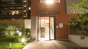 Entrada a un edificio con puerta de vidrio, un pequeño jardín con sendero y una escalera que da acceso al interior.
