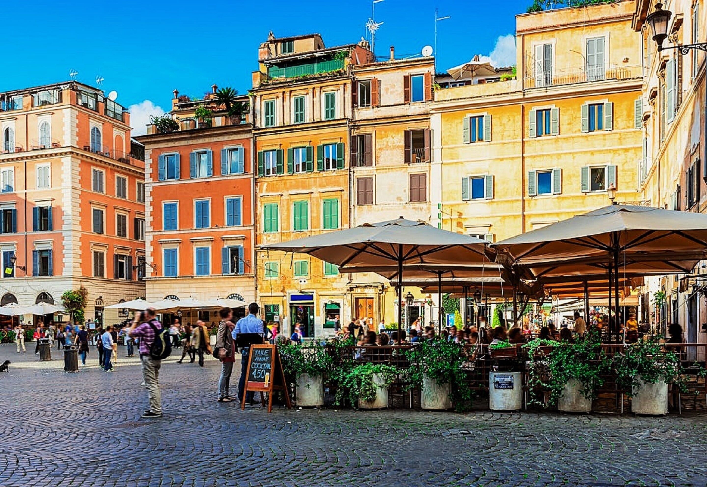 A cobblestone square with outdoor cafes, historic buildings, and a signboard.
