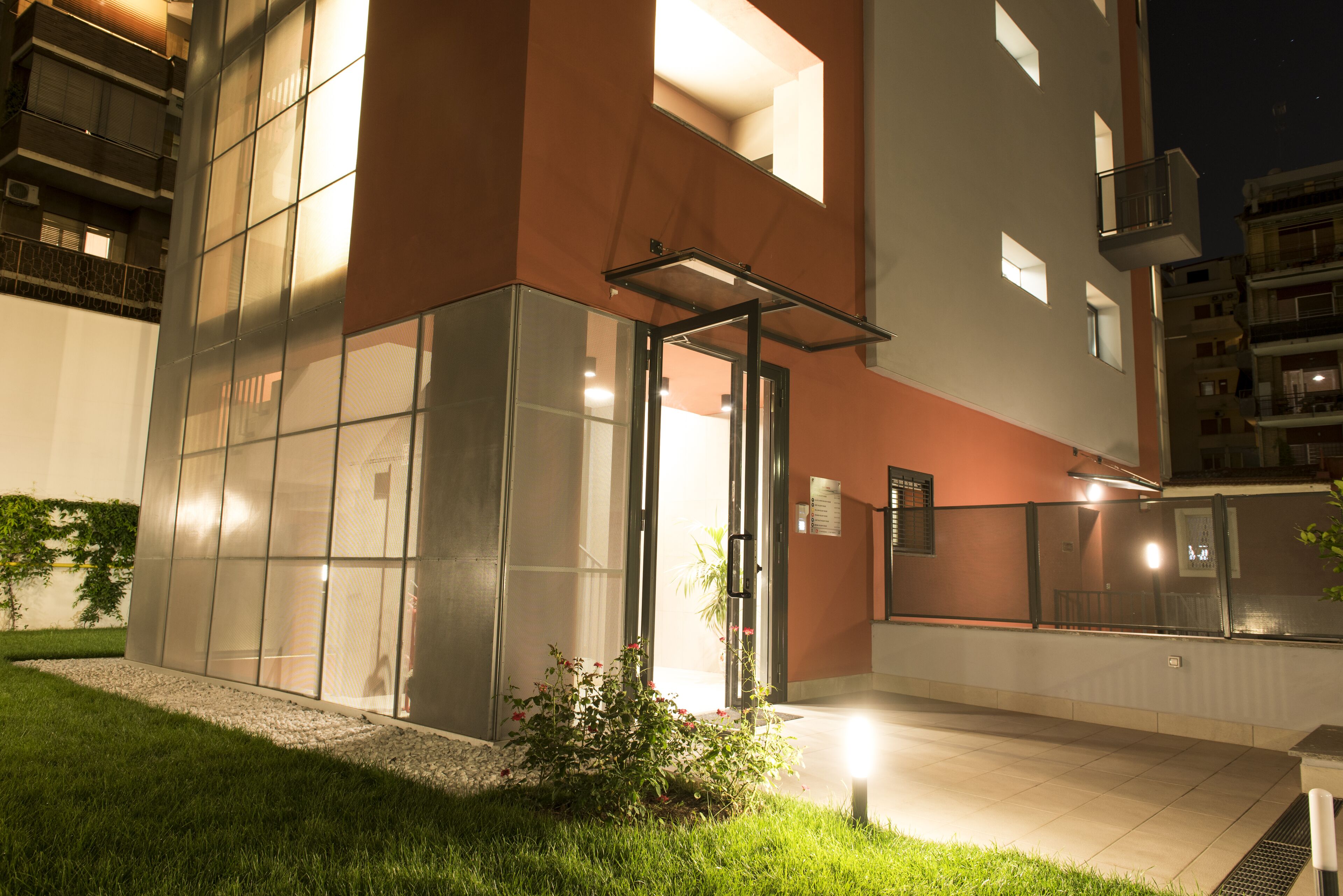 A modern building with a glass entrance, a small garden area, and a well-lit exterior.