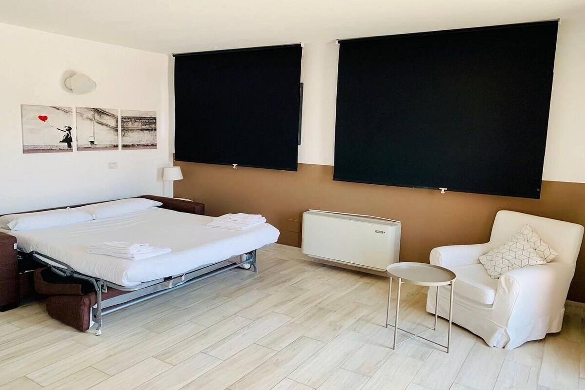 Habitación de hotel con cama, una silla, una mesita y dos cortinas negras grandes.