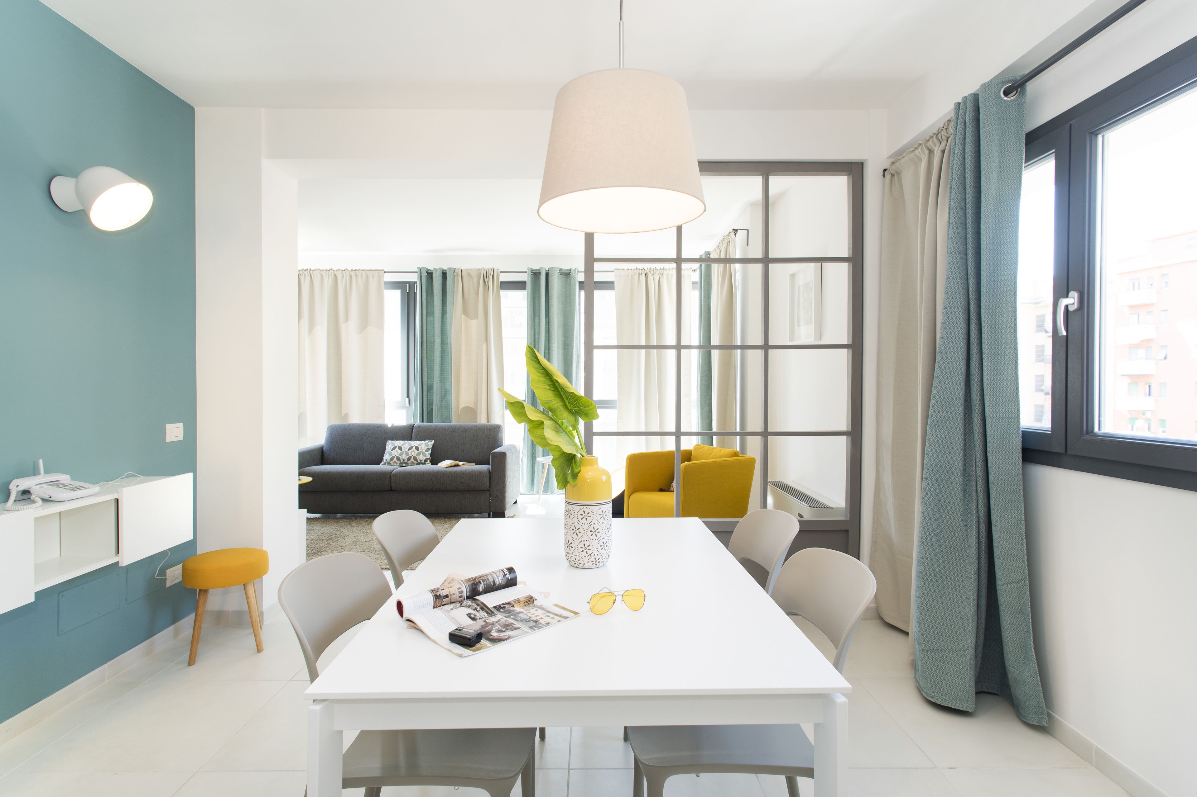 A modern living room with a sofa, a dining table with chairs, a pendant light, and a large window with curtains.