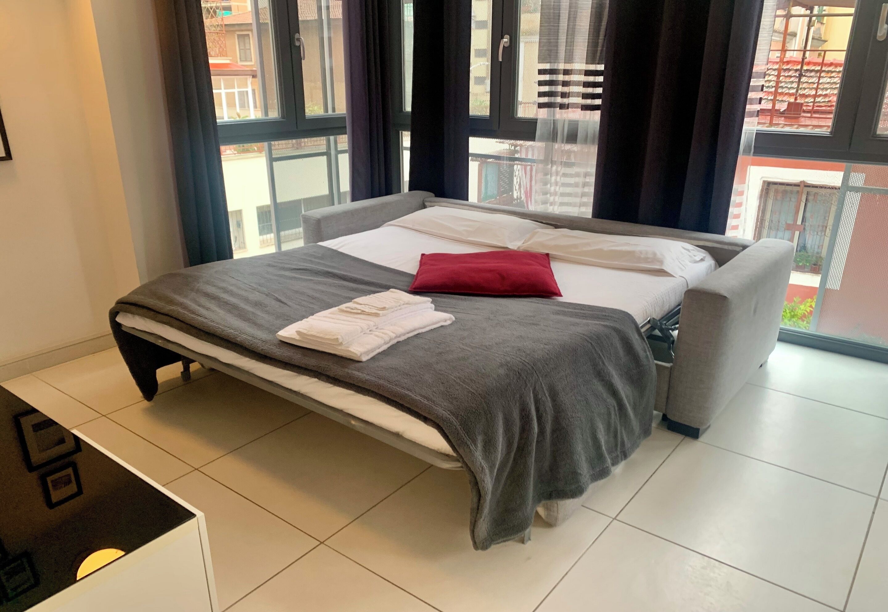 A neatly made bed with a grey blanket and a red pillow, a white towel, and a grey upholstered armchair.