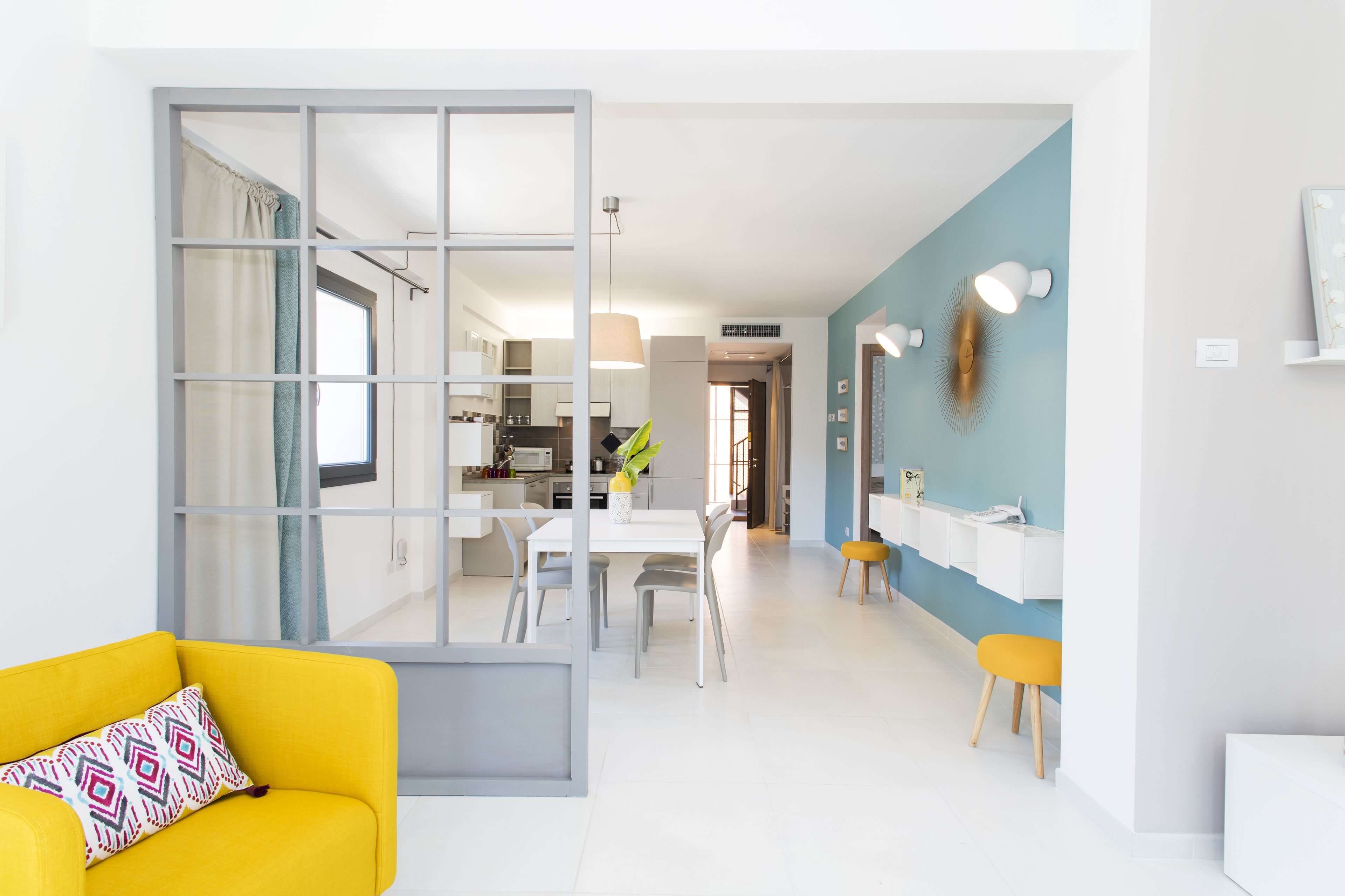 A modern living room with a yellow sofa, a dining area with a white table and chairs, and a kitchen visible through a glass partition.