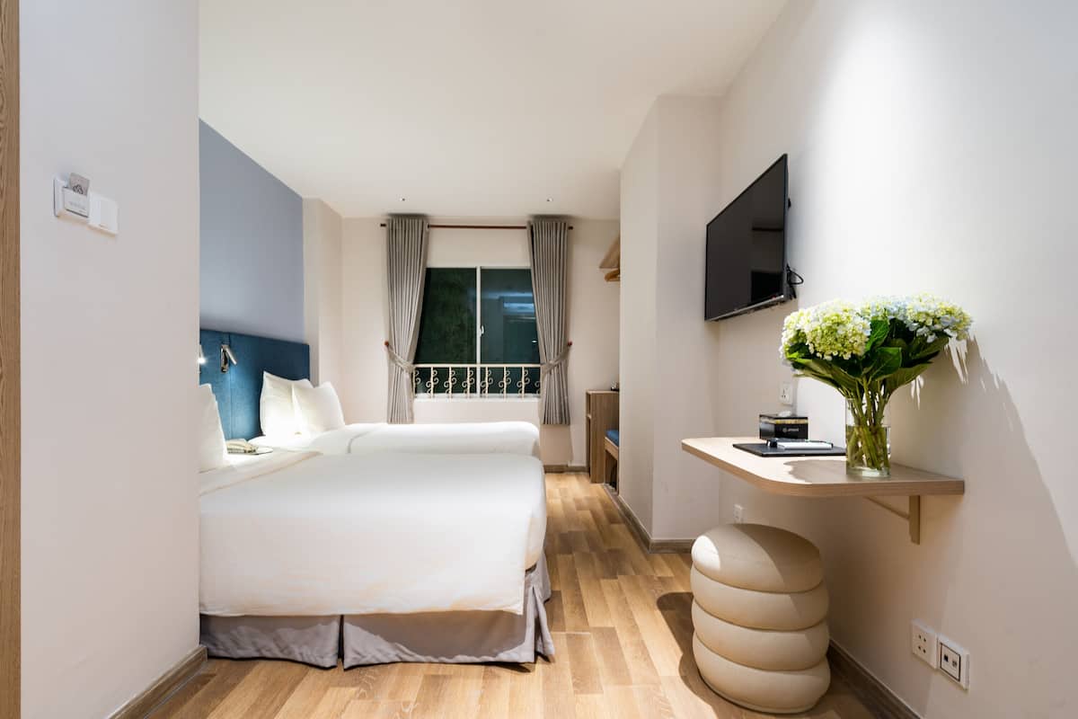 Hotellihuone, jossa on sänky, seinälle asennettu televisio, pieni työpöytä, jossa on maljakko täynnä kukkia, ja ikkuna, jossa on verhot.