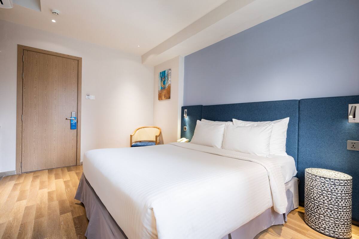 Hotellihuone, jossa on suuri sänky, puinen ovi, sininen tehosteseinä ja kuvioitu sivupöytä.