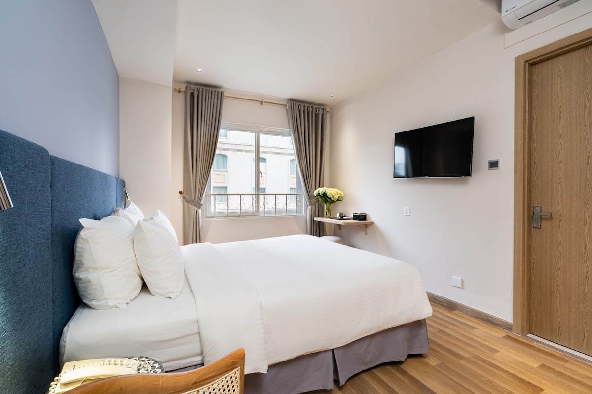 Hotellihuone, jossa on sänky, televisio, ikkuna verhoilla, puuovi ja pieni pöytä, jonka päällä on maljakko.