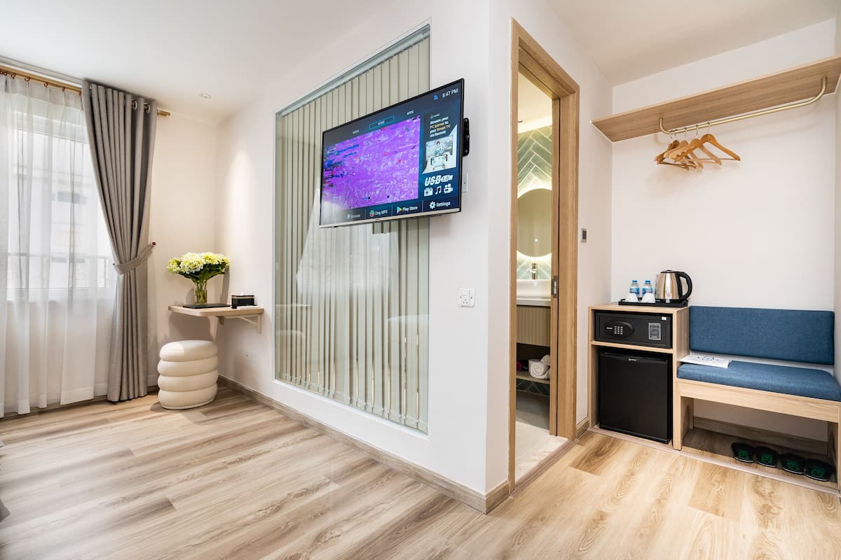 Moderni hotellihuone, jossa on taulutelevisio, sohva, pieni keittonurkkaus ja kenkäteline.