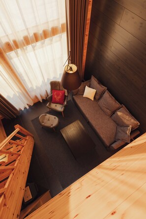 A room with a wooden floor, a sofa with cushions, a chair with a red cushion, a small table, and a hanging lamp.