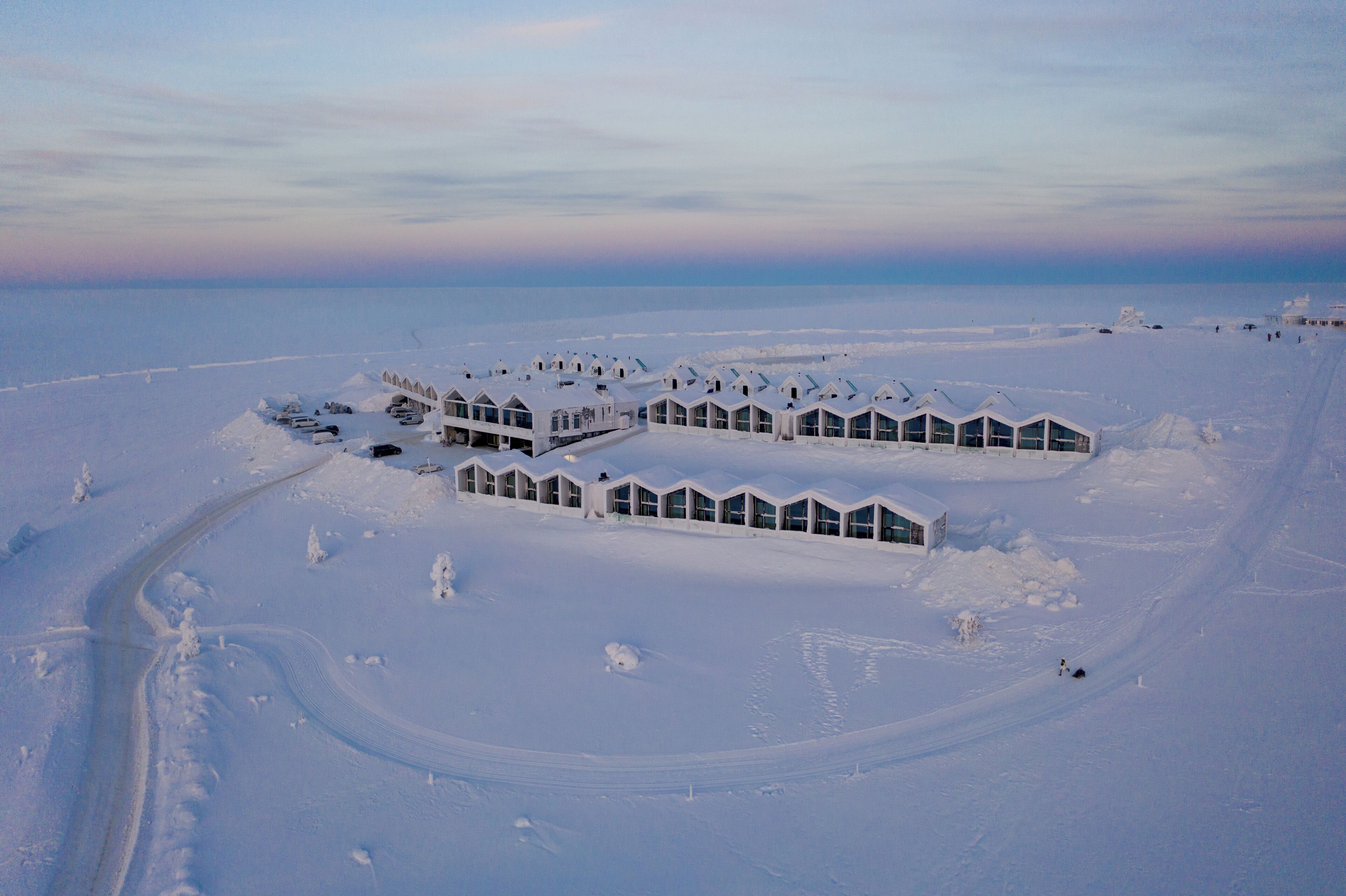 A snow-covered resort with multiple buildings, a winding road, and a clear sky.