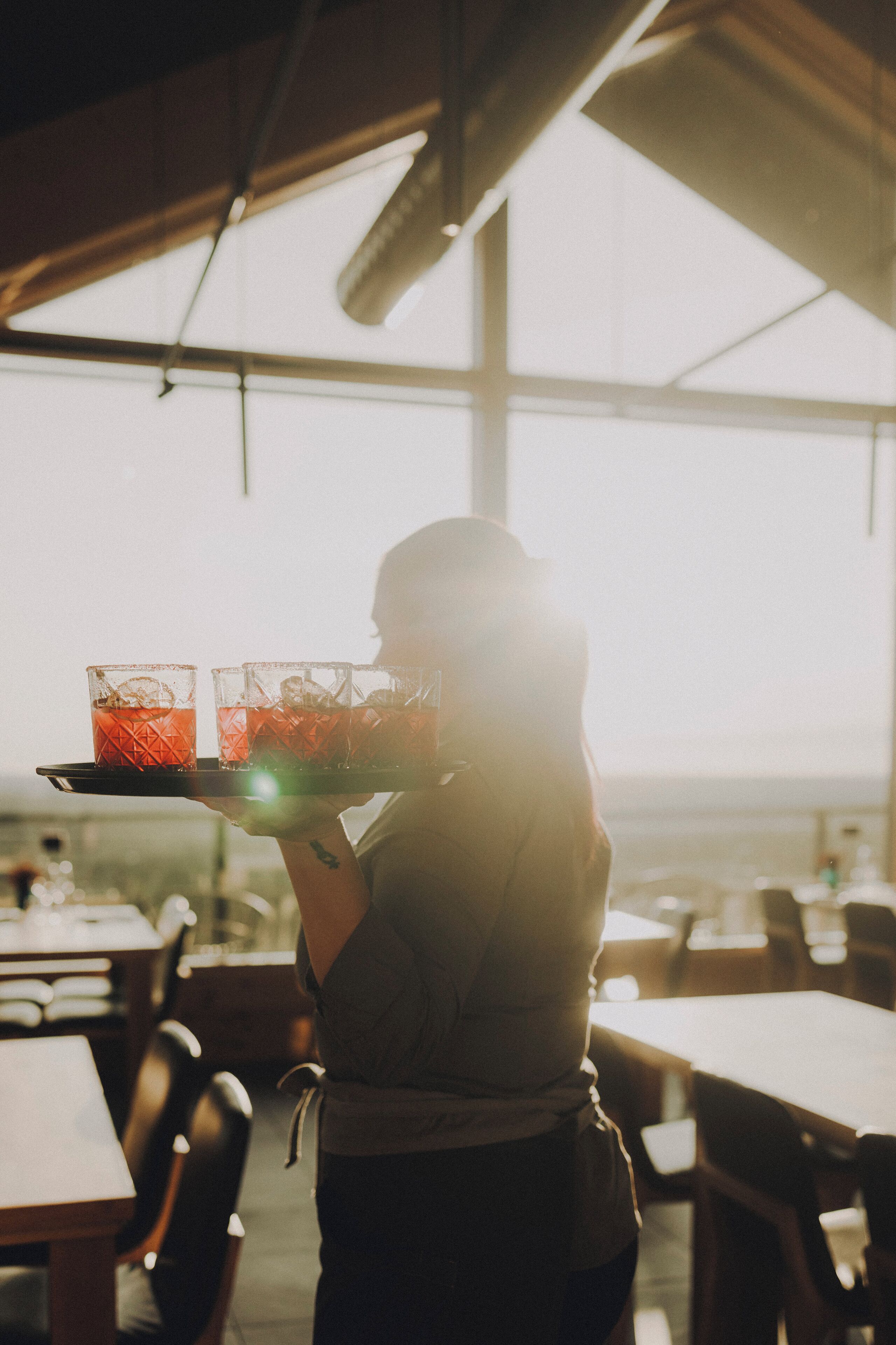 A person holding a tray with two glasses of red drink, in a well-lit indoor setting with tables and chairs.