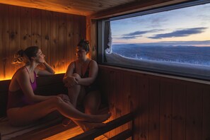 Two individuals seated by a window in a wooden cabin with a view of a snowy landscape.
