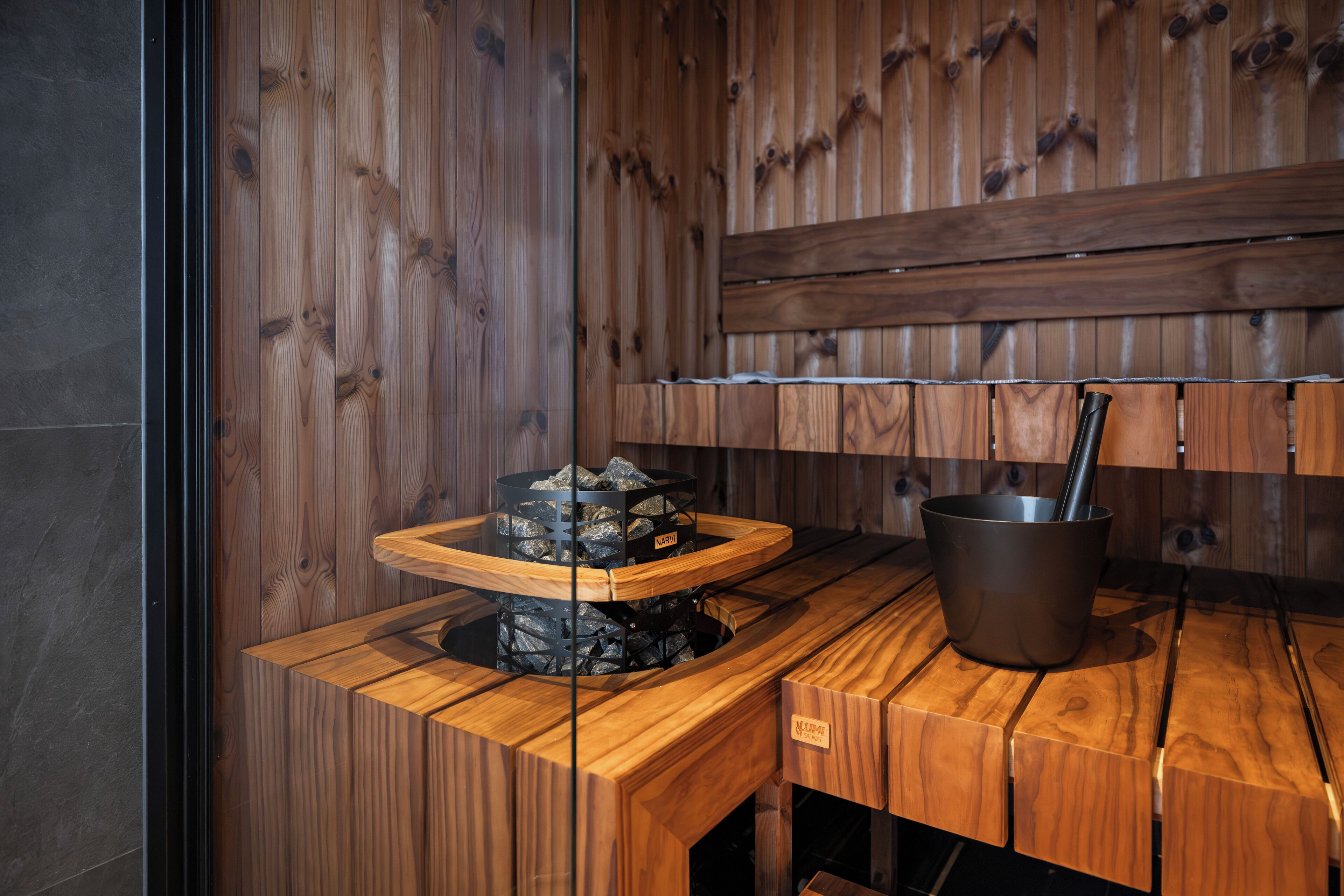 Una sauna de madera con un banco de piedra, un cubo de metal y un estante de madera.