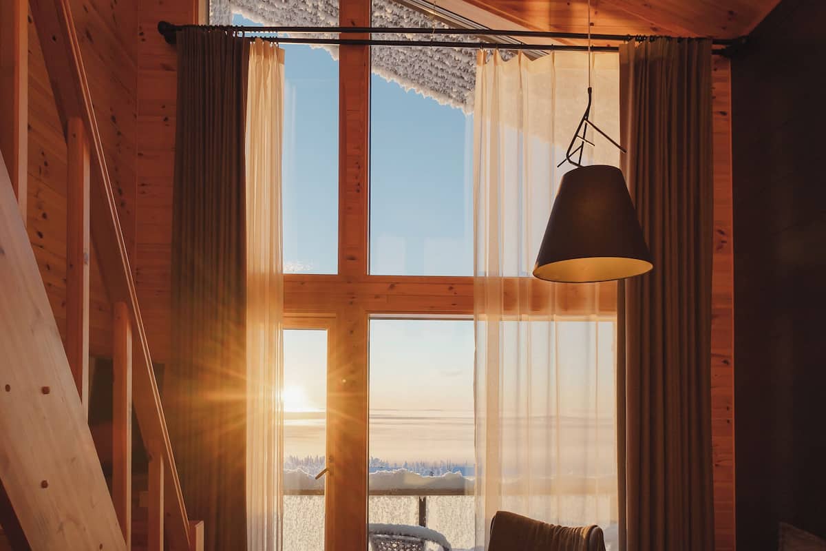 A cozy cabin interior with a wooden staircase, a sofa, a chair, and a large window offering a view of the outdoors.