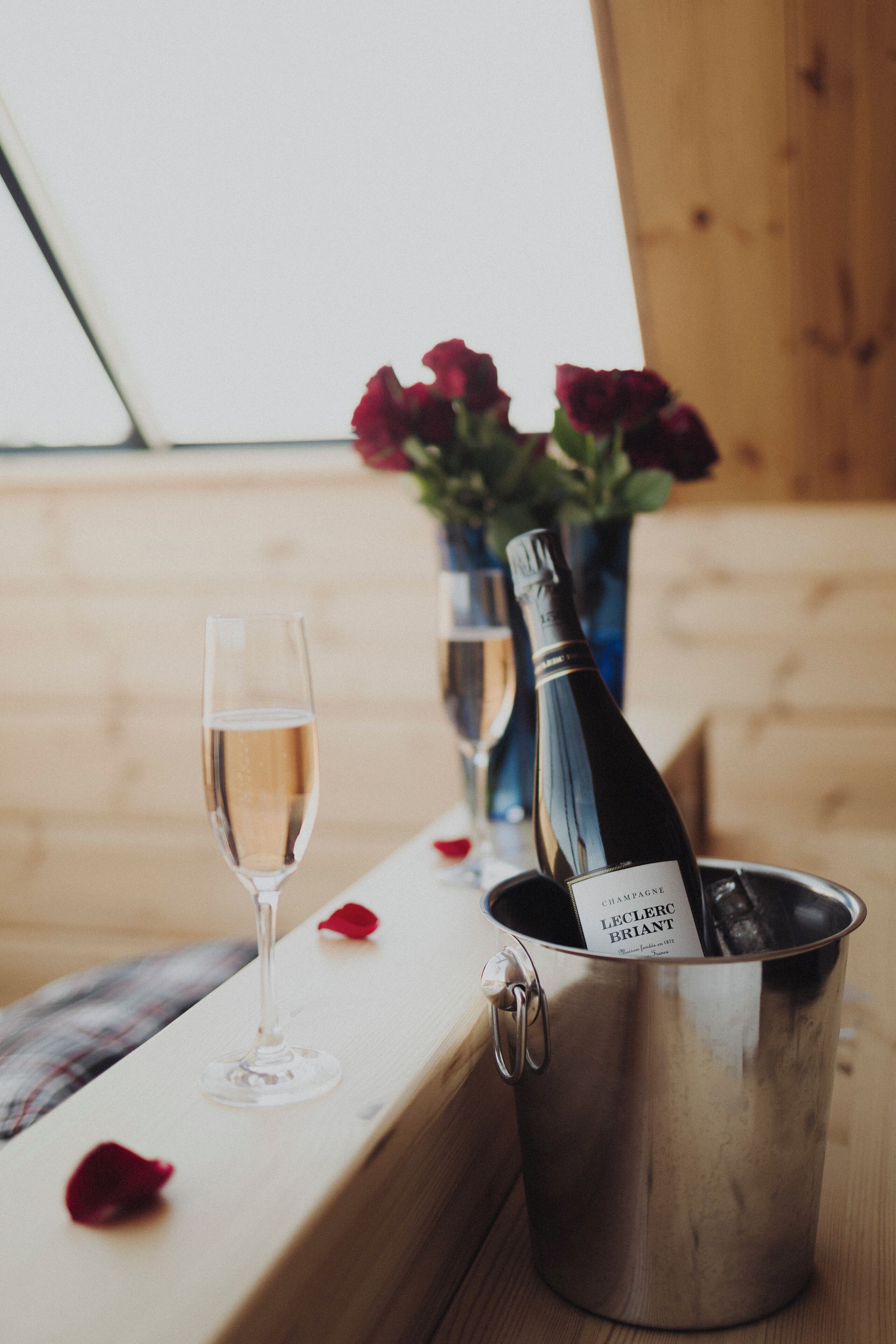 Una bottiglia di champagne in un secchio di metallo, un bicchiere di champagne e un vaso con fiori rossi su un tavolo di legno.