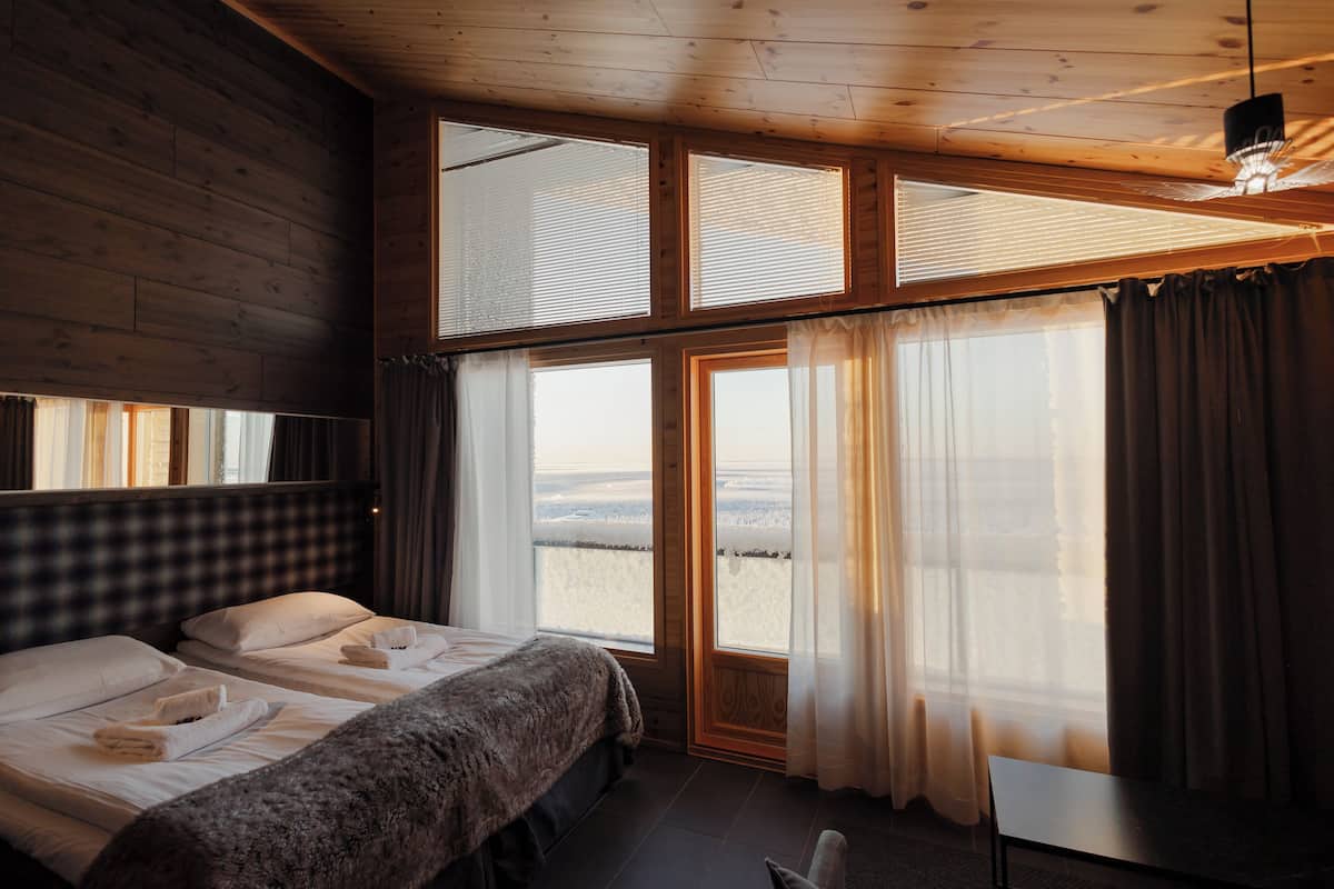 A room with two beds, large windows with curtains, and a view of a snowy landscape.