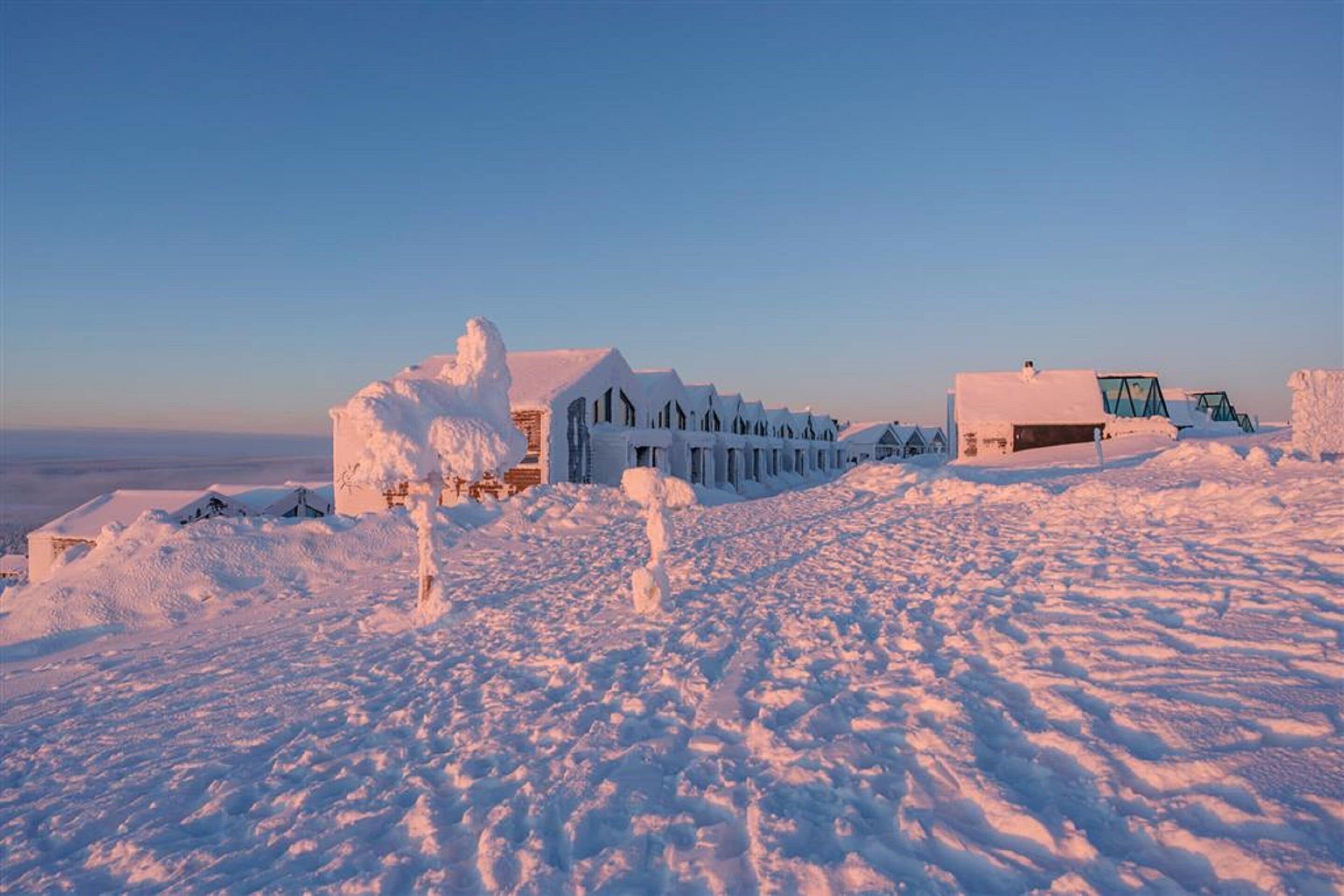 A snow-covered landscape with buildings, including a large house and smaller structures, under a clear sky.
