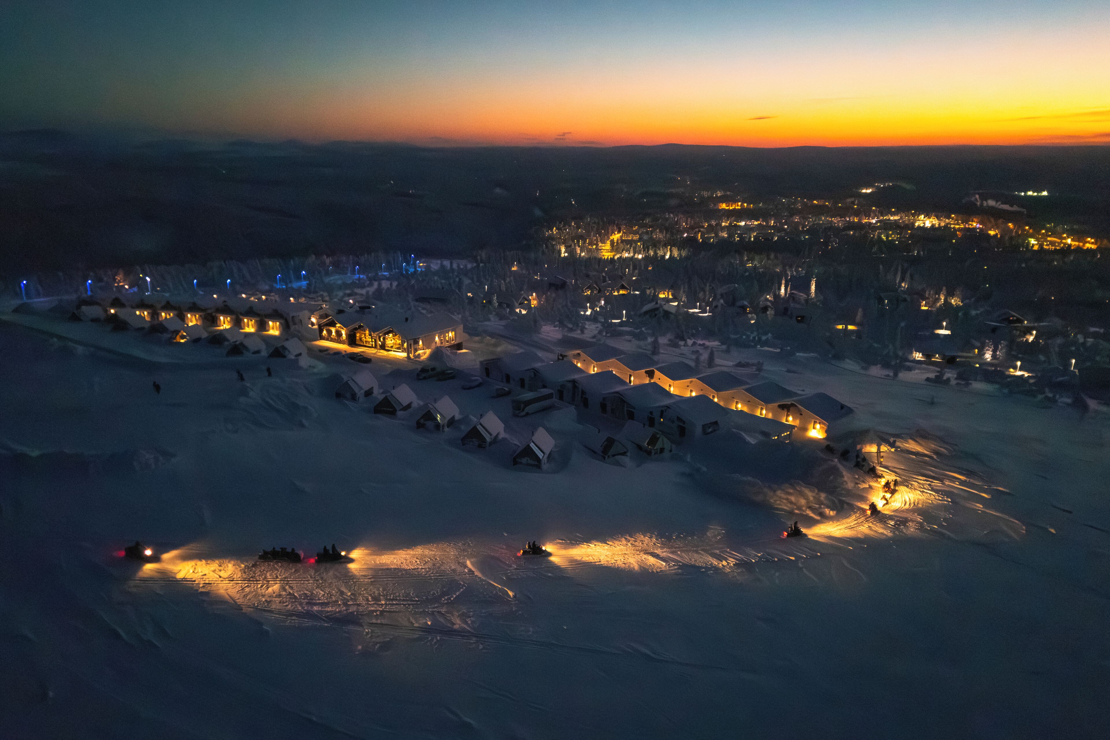 Schneebedeckte Landschaft mit mehreren Holzhütten und einem beleuchteten Bereich im Vordergrund.