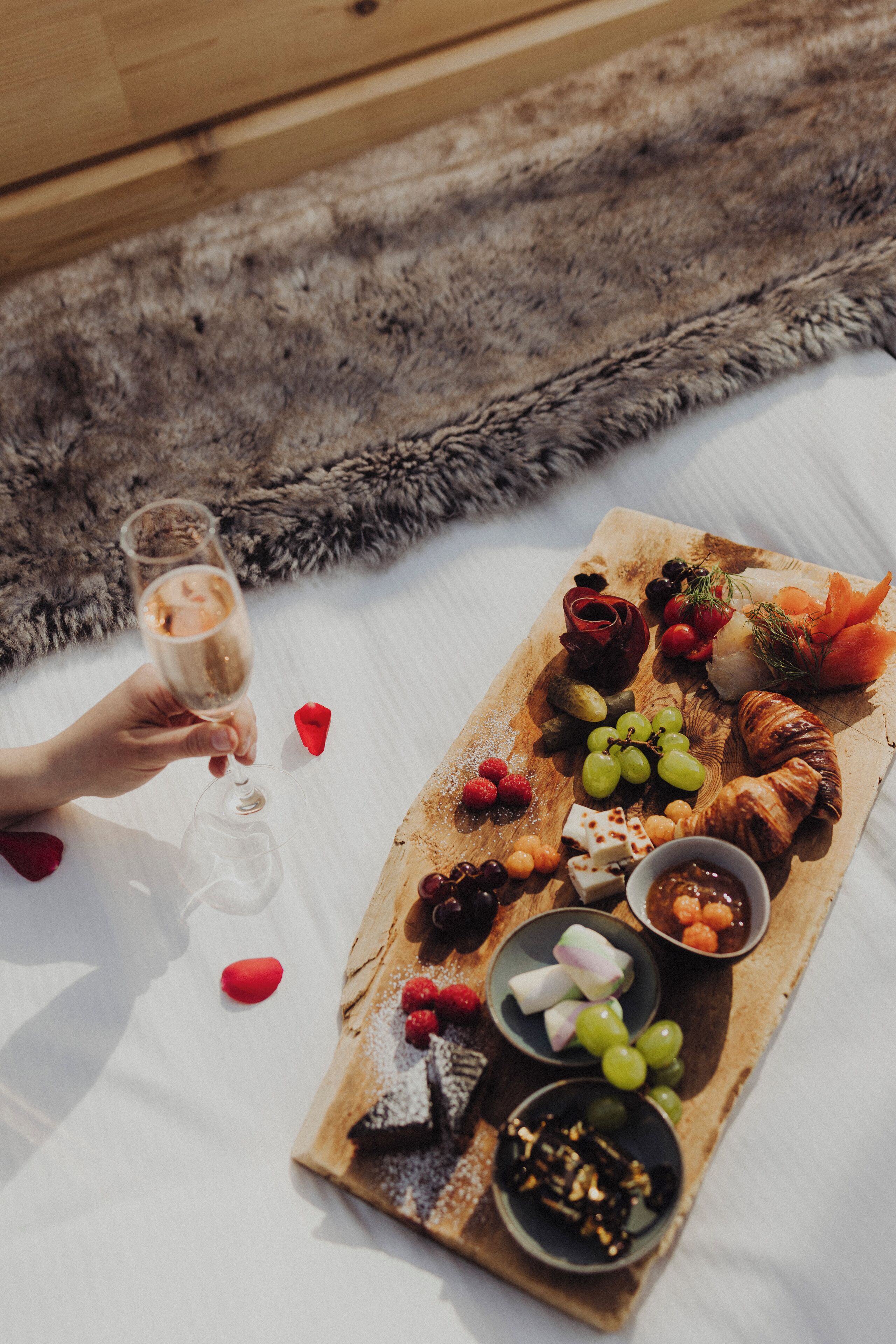 En picknickuppsättning med en träbricka som innehåller olika sorters mat och dryck, inklusive vindruvor, ost och en croissant, placerad på en vit yta.