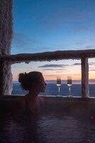 Una persona en un jacuzzi, con dos vasos de agua, una ventana que ofrece vistas al cielo y montañas.