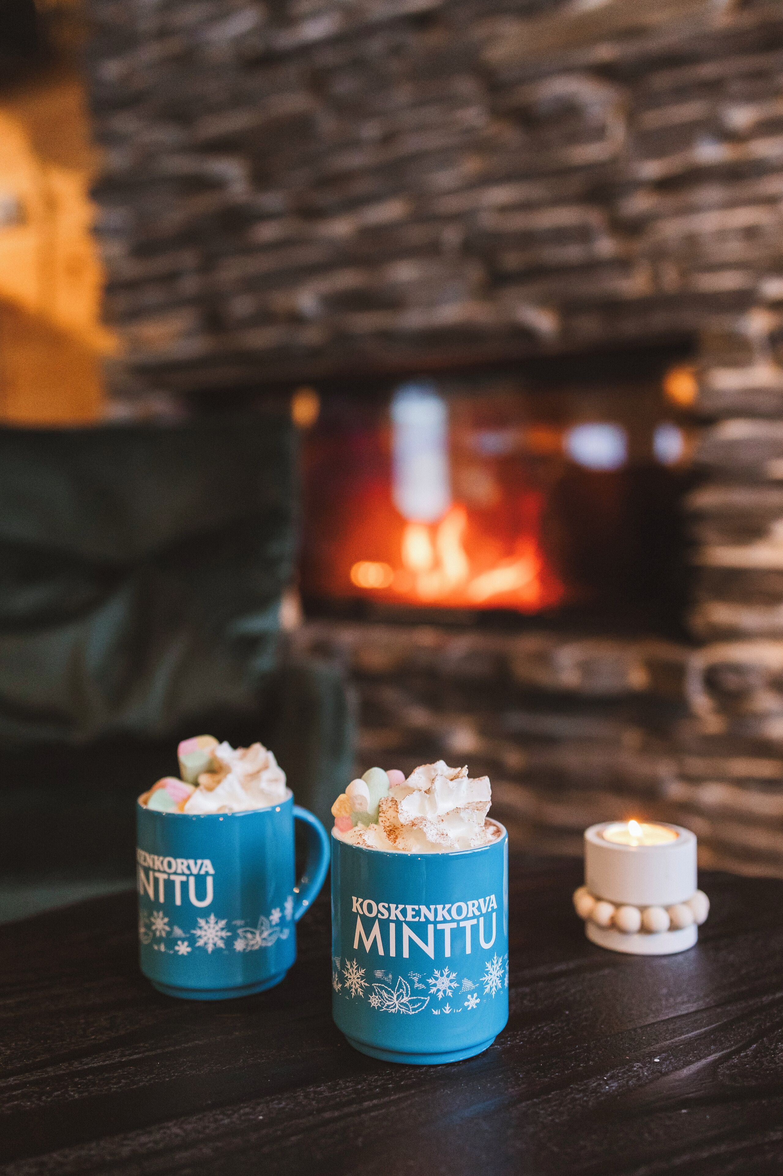 Deux tasses bleues ornées de motifs en forme de flocons de neige et d’inscriptions blanches, posées sur une surface en bois sombre, avec une bougie allumée et une cheminée en arrière-plan.