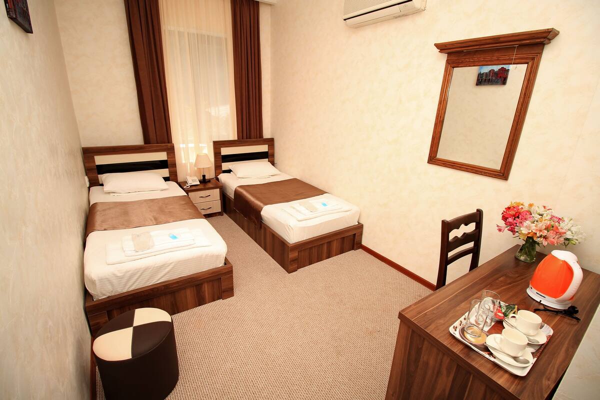 Habitación de hotel con dos camas, un escritorio con silla, una mesita, un florero con flores y un equipo de aire acondicionado.