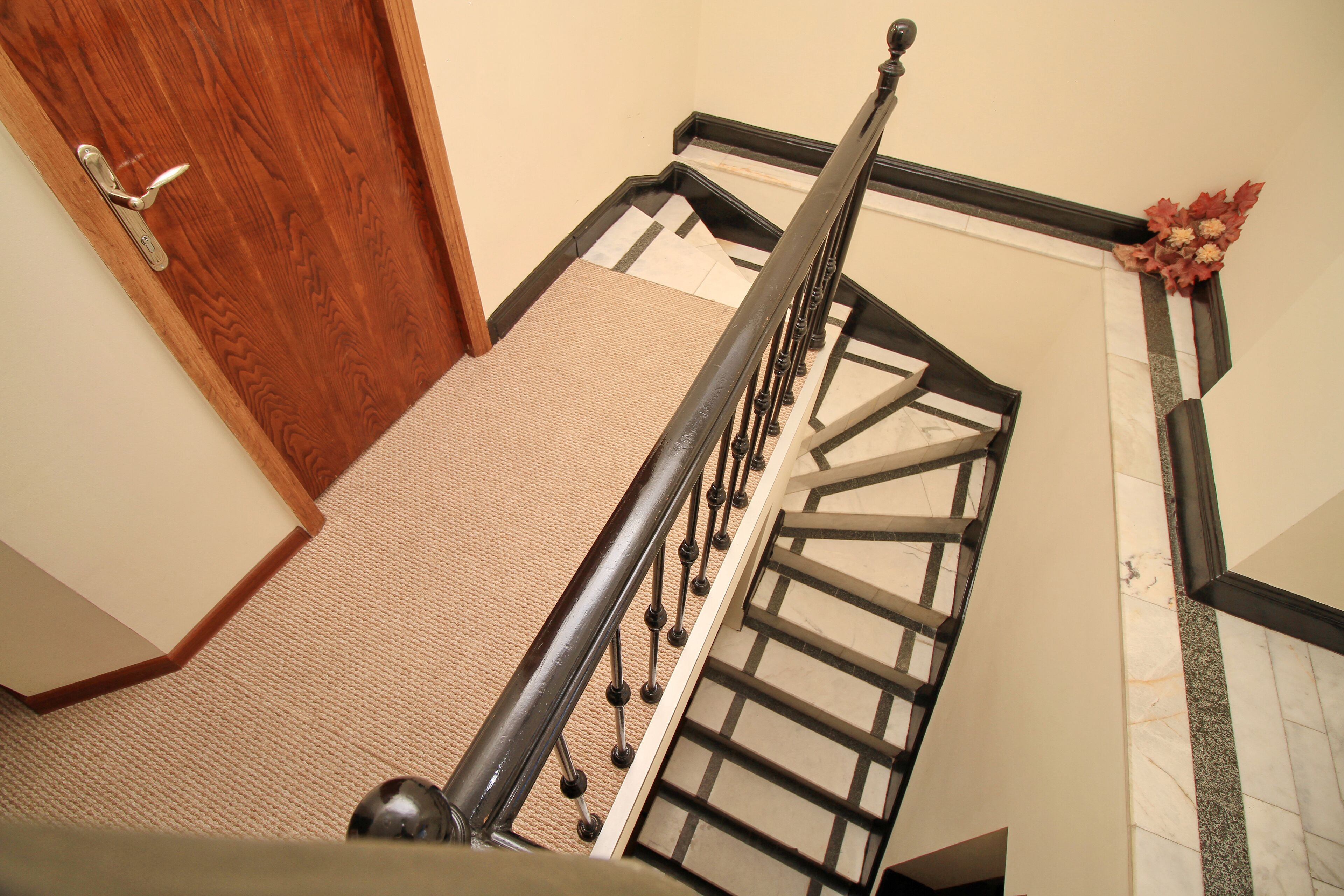 A wooden door with a silver handle, a carpeted staircase with a black metal railing, and a wall with a white baseboard and a decorative floral arrangement.