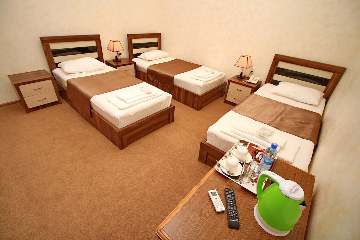Habitación de hotel con cuatro camas individuales, una mesita de noche de madera con una mesita abatible, y una pequeña mesa con una tetera y controles remotos.