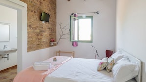 A bedroom with a brick wall, wooden beams, a bed with white linens, a bedside table, a TV, and a window with a view. - Hostal Cling 43 (Deltebre)