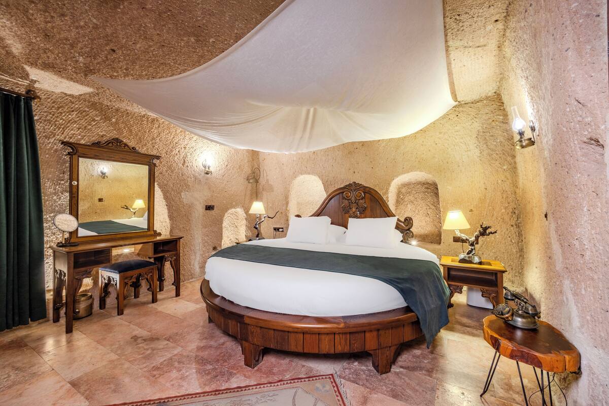 A circular wooden bed with white bedding, a mirror, a small table, and a lamp on a stone wall.