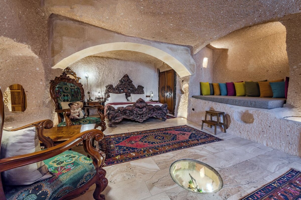 A room with a stone wall, a wooden chair, a bed with a patterned rug, and a shelf with colorful cushions.