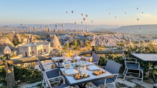 Un espace de restauration en plein air avec une table dressée pour deux personnes, offrant une vue sur un paysage composé de formations rocheuses uniques et, au loin, sur des montgolfières.