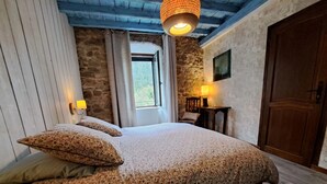 A bedroom with a large bed, stone walls, wooden ceiling beams, a wooden door, and a window with curtains. - Le Sareymond (Chatillon-en-Diois)