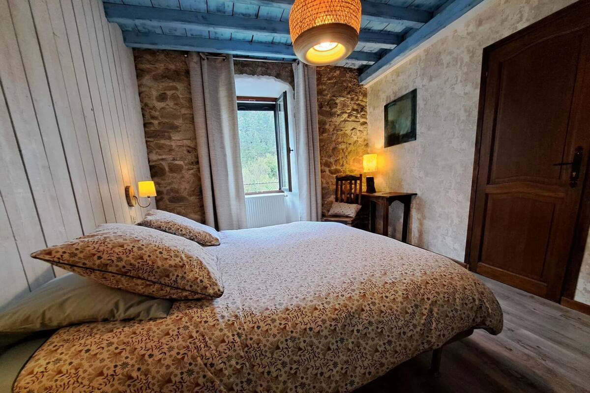 A bedroom with a large bed, stone walls, wooden ceiling beams, a wooden door, and a window with curtains.