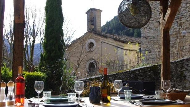 A dining table set outdoors with wine bottles and glasses, overlooking a stone building and trees. - Le Sareymond (Chatillon-en-Diois)